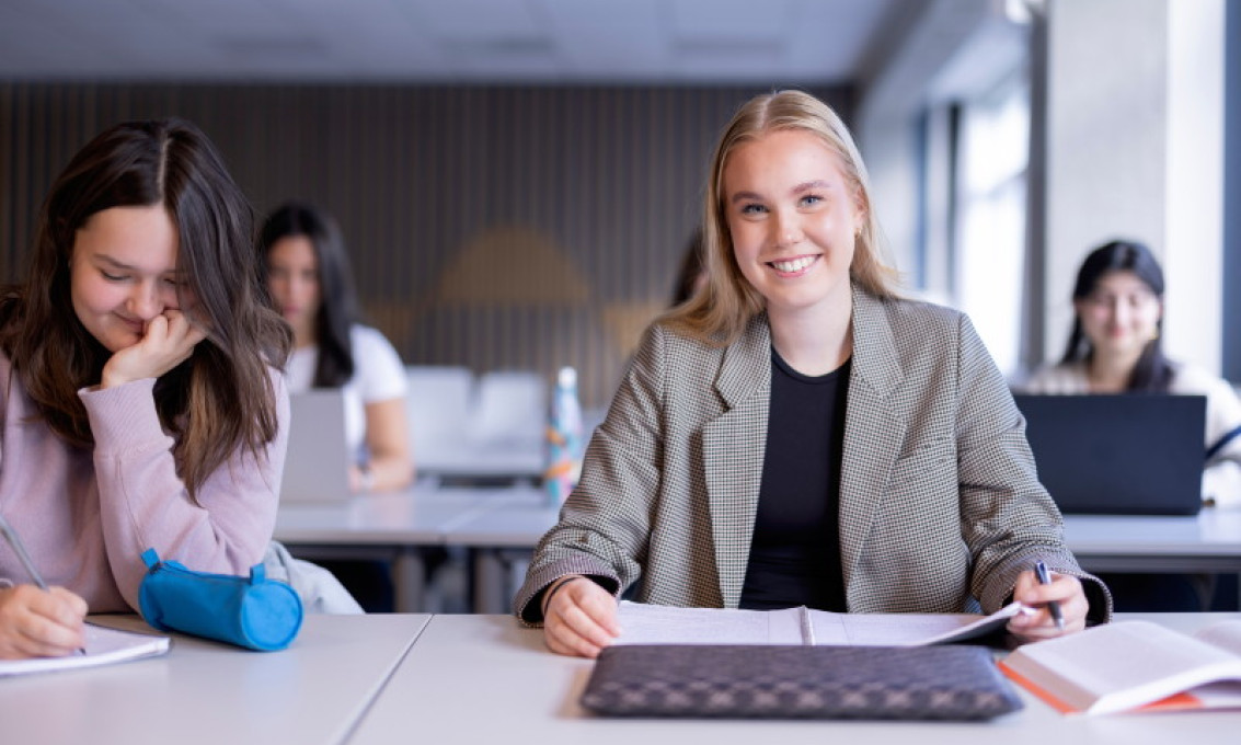 To kvinnelige studenter sitter ved et skrivebord i et forelesningsrom. Den ene smiler mot kamera. To andre studenter ses i bakgrunnen.