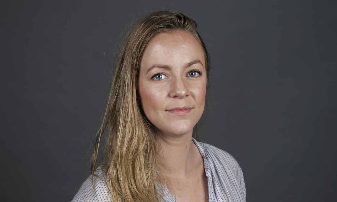 Kvinne i halvfigur ser inn i kamera. Mørk bakgrunn.
