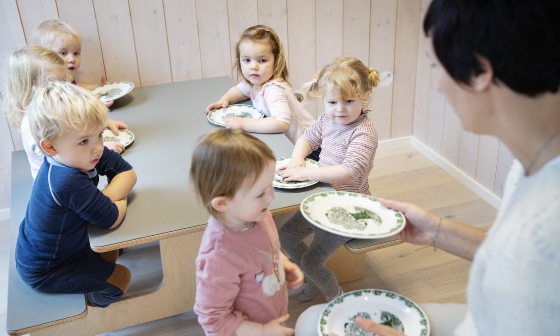 Fem barn rundt et barn i barnehagen. Ett barn får utdelt en tallerken av en barnehagelærer