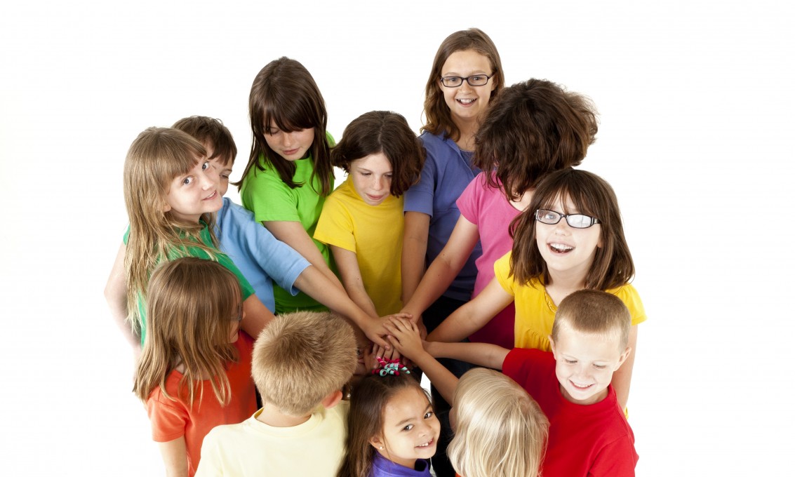En gruppe barn i forskjellig alder står i en sirkel med en hånd hver inn mot midten. Foto: iStock.
