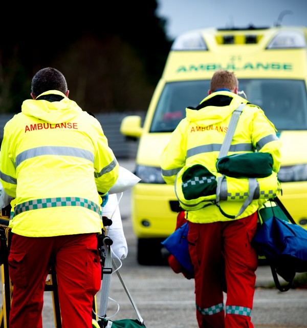 To ambulansepersonell i uniform frakter pasient på båre til ambulansen