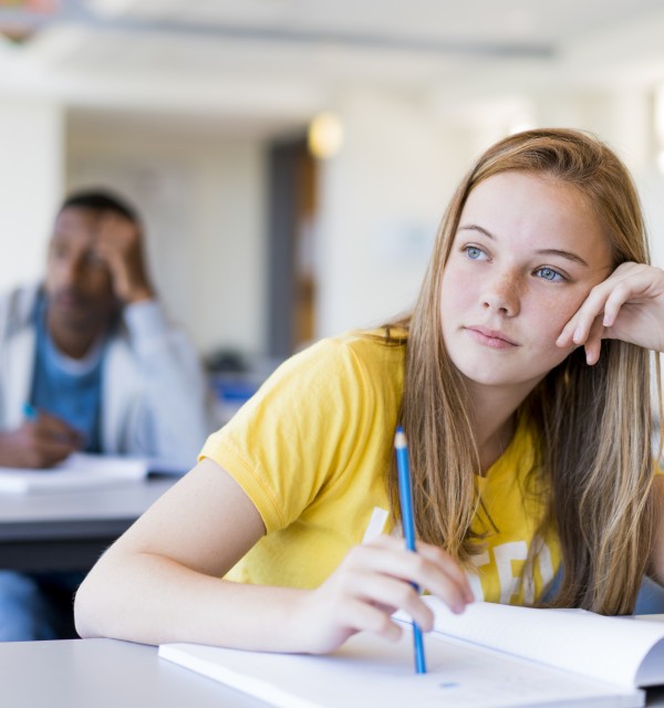 Jente sitter ved pulten sin i et klasserom med en skrivebok foran seg og pennen i hånden. Det ser ut som om hun tenker på noe
