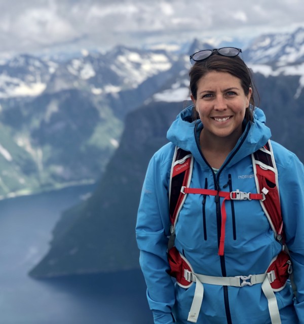 Ung kvinne er på fjelltur og står i blått turtøy på en høy topp og smiler til fotografen, med fjell og fjord i bakgrunnen.