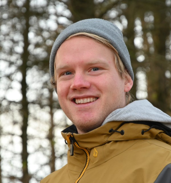 En ung mann med grå lue og gul jgakke med hette står i skogen og smiler til kamera
