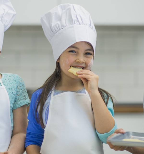 Tre barneskoleelever er på skolekjøkken ikledd forkle og hvite kokkehatter, de smaker på småkaker.