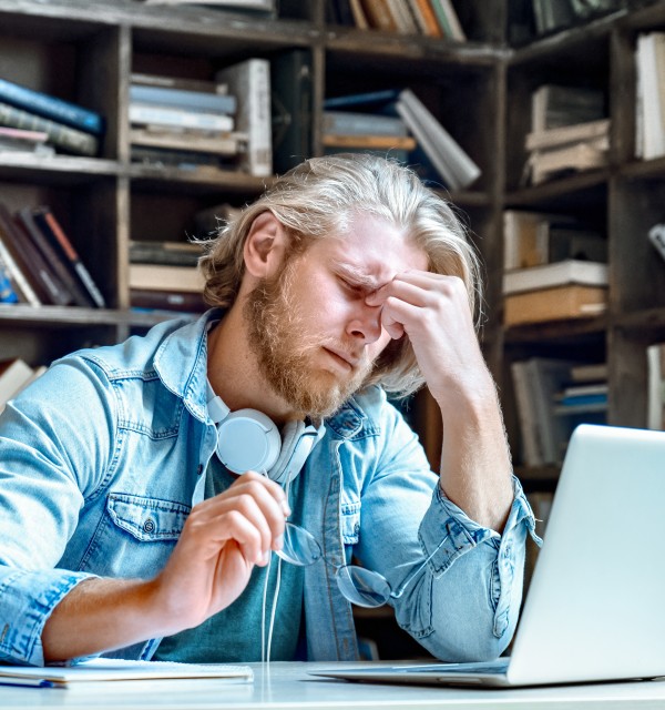 En mann sitter ved datamaskinen med bøker rundt seg. Han holder fingrene mot panna og ser sliten ut. 