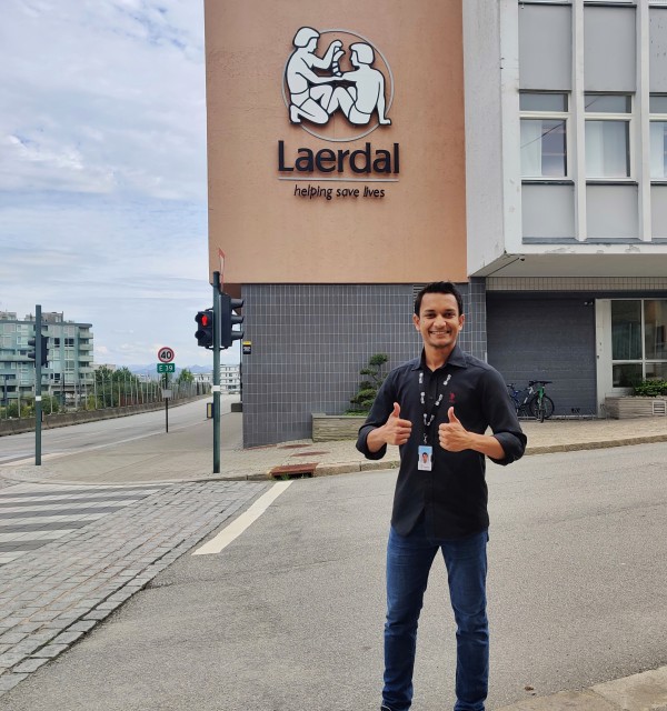 Navid står fremfor inngangspartiet til Laerdal Medical, han smiler til kamera og holder to tomler opp 