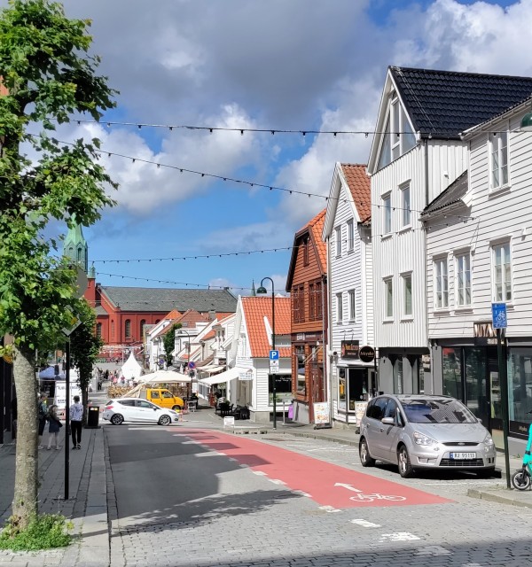 Pedersgata i Stavanger med hvite og oransje hus på høyre side og en rød bygning på venstre side. Et grønt tre står på venstre side, og en grå bil er parkert på høyre side. En mann går på høyre fortau. Foto.