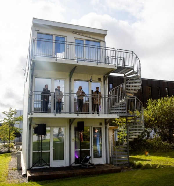 Fire personer står på en balkong i andre etasje på et tre etasjers bygg. Foto. 