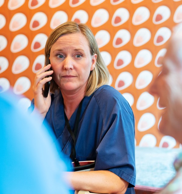 Hjemmesykepleier i blå uniform snakker i telefon mens hun ser på pasienten sin