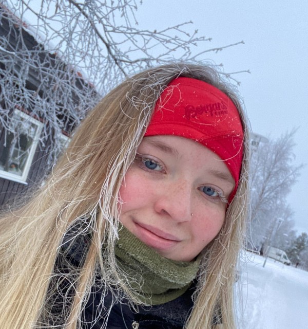 ung kvinne med langt, lyst hår og rødt pannebånd i vinterlig, hvitt landskap. Hun har snø i håret og frost på øyevippene.