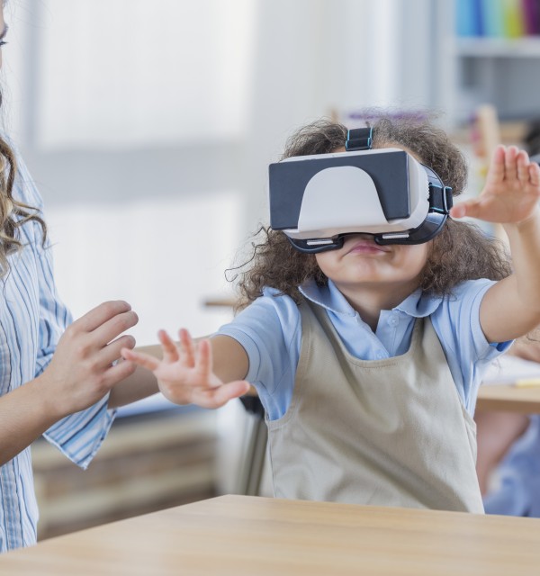 førskolebarn holder armene foran seg i luften, har VR-briller på hodet. Ved siden av barnet sitter en voksen kvinne og følger med på barnet. Hun smiler og holder hendene sine ut mot barnet.