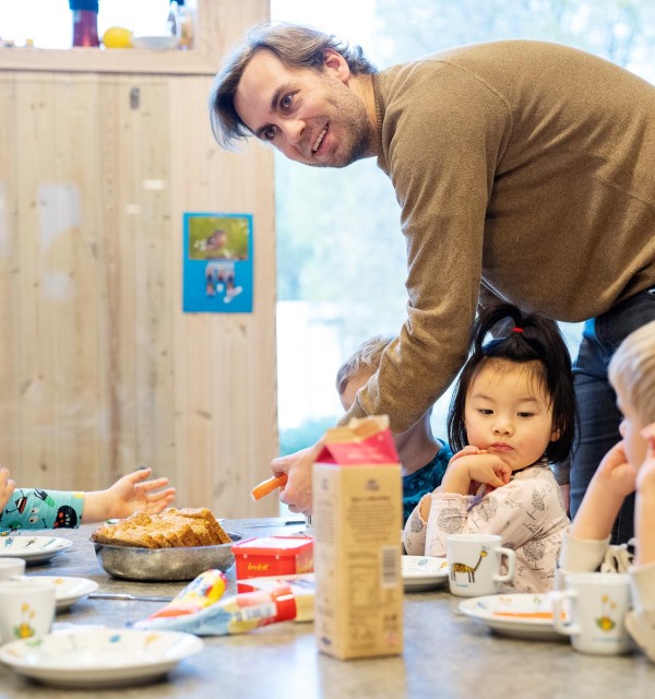 mann står bøyd over barn som sitter og spiser ved et lavt bord