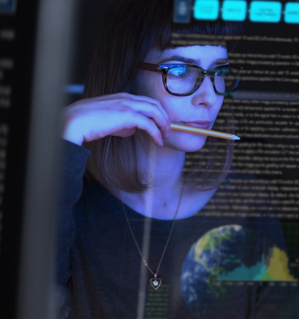 kvinne har blyant mellom tennene og ser på dataskjermer, omgitt av mørke, lyset fra skjermen lyser opp ansiktet hennes