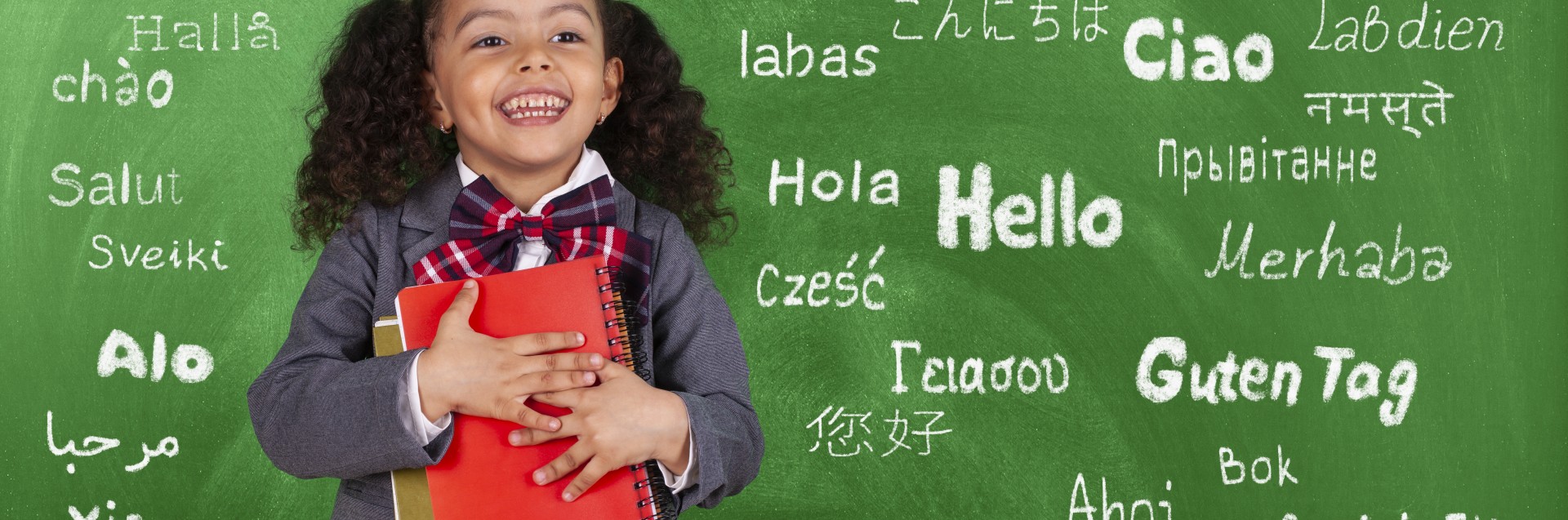 En jente står foran en tavle med en bok i armene. På tavlen står det "hallo" på flere språk