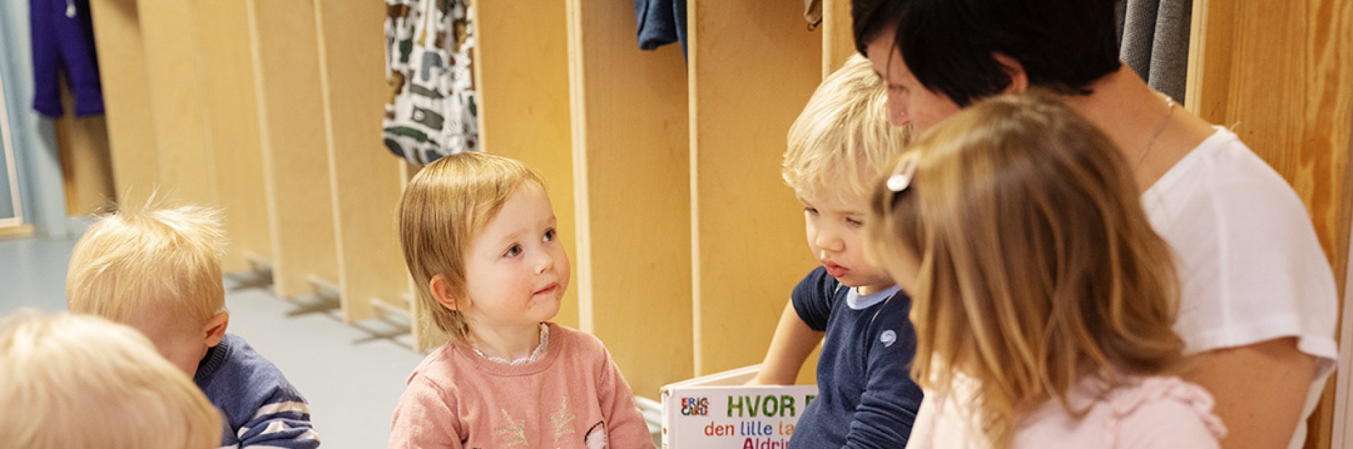 Barnehageansatt sitter i garderoben og leser bok for seks barn. Har god blikkontakt med et av barna. To andre barn sitter på fanget. De andre sitter rolig rundt henne i en sirkel.