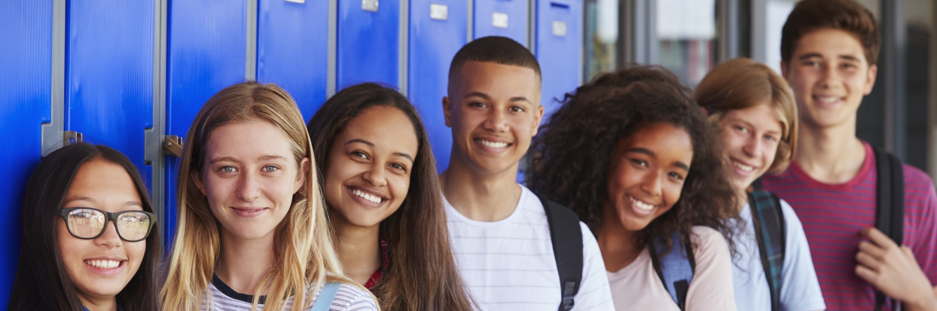 En gruppe smilende ungdommer står etter hverandre ved en skaprekke på en skole.