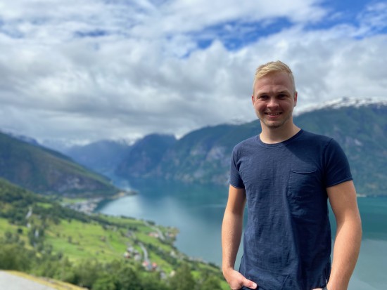 Mann med lyst hår står ute i naturen med fjord og fjell i bakgrunnen