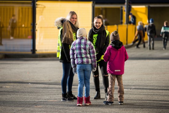 To kvinnelige lærerstudenter står utendørs sammen med to elever med ryggen til i et friminutt på en skole.