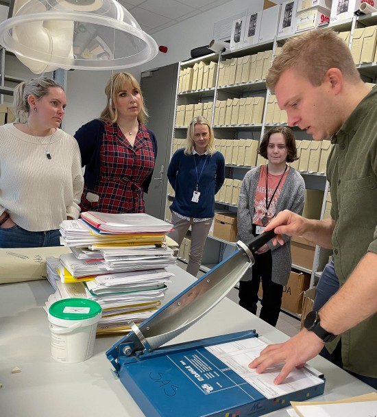 En gruppe mennesker ser på en mann som skjærer etiketter i et arkiv