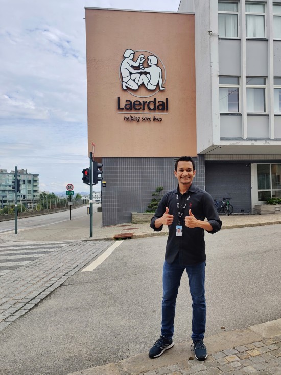 Navid står fremfor inngangspartiet til Laerdal Medical, han smiler til kamera og holder to tomler opp
