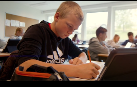 Nærbilde av gutt i klasserom som skriver på ark med chromebook foran seg på pulten. Andre elever i bakgrunnen.