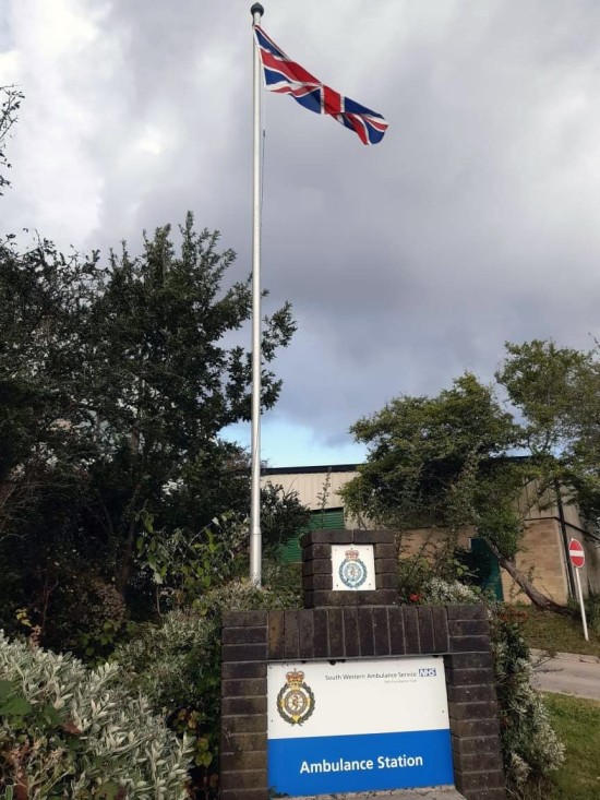 BIldet viser skiltet til ambulansestasjonen under en flaggstang med britisk flagg.