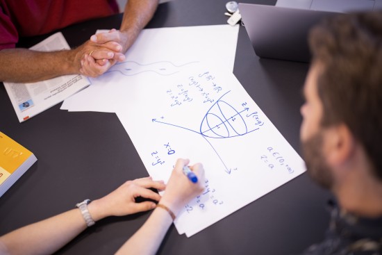 Tre personer sitter rundt et bord og ser på  et ark med tegning av matematiske modelleringer på.