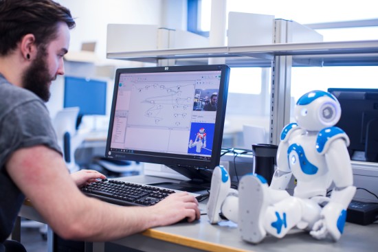 En mann sitter foran en datamaskin. Ved siden av sitter en liten hvit og blå robot.