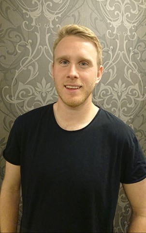 Mann med lyst hår og svart t-skjorte ser på kamera.