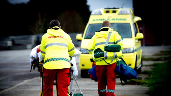 To ambulansepersonell i uniform frakter pasient på båre til ambulansen