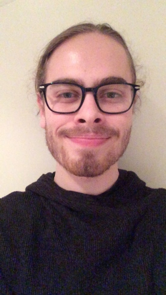 Ung mann med svarte briller og langt hår trukket i hestehale har på seg svart hettegenser og smiler med lukket munn til fotografen. Bakgrunnen er en hvit vegg.