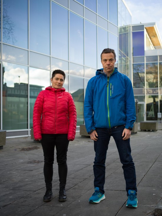 Kvinne med rød jakke og mann med blå jakke står oppreist utendørs og ser alvorlig rett inn i kamera