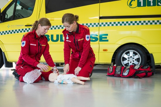 To kvinnelige paramedisinere i rød uniform gjør livredning på en babydukke foran en ambulanse.