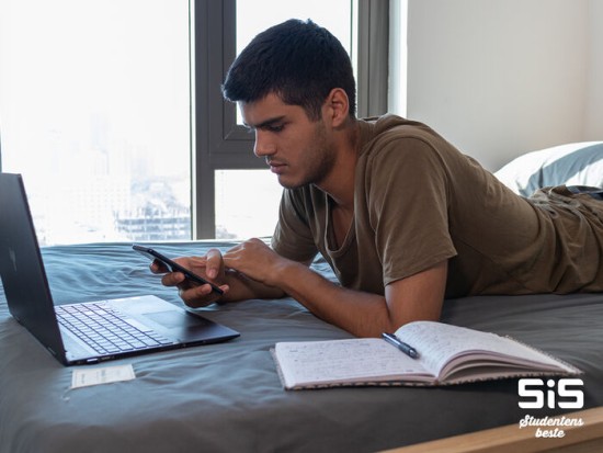 Student ligger på sengen og ser på mobiltelefon samtidig som han ser på laptop og leser bok