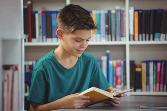 ung gutt i grønn t-skjorte leser i en bok. bokhylle i bakgrunnen.