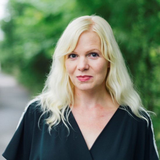 Portrett av kvinne med blondt hår og sort topp. Grøntområde i bakgrunnen.