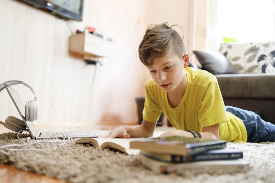 Ung gutt i gul t-skjorte ligger på gulvet i en stue og leser bok. Det ligger tre bøker ved siden av ham på gulvet, sammen med en pc og hodetelefoner.