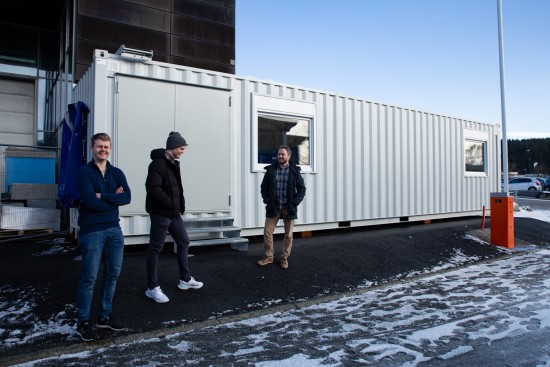 Tre menn står foran en container som inneholder en vindtunnel.