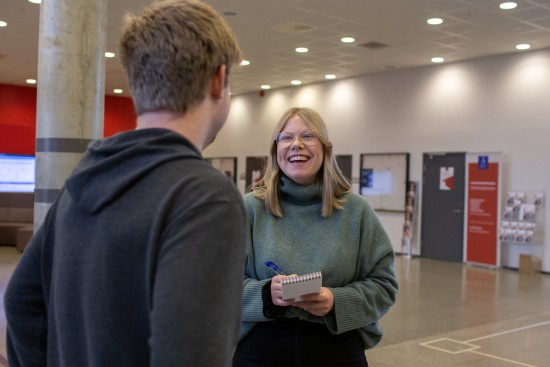 Bilde av en student med notatbok og penn som smiler til en annen person som står mer ryggen mot kameraet.
