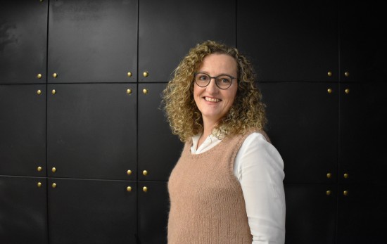 Professor i barnevern og nettverksleder Ingunn T. Ellingsen. Hun og er iført en hvit skjorte med beige vest. Hun har briller og mørkeblondt krøllet hår, hun står vendt til siden, vekk fra kameraet med kroppen og ser inn i kameraet. Bak henne er en mørk vegg med gulldetaljer.