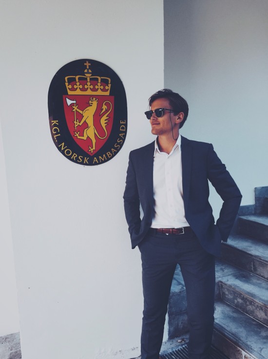 Med dress og solbriller inne i den norske ambassaden i København. Johannes står foran en vegg  med bilde av det norske våpenskjoldet bak han: en sort oval med en rød trekant med krone oppå og en løve med en øks. 