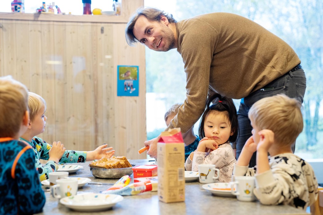 Mannlig barnehageansatt hjelper en gruppe barn som sitter ved et bord og spiser