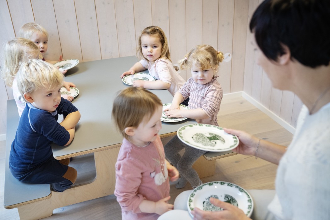 Fem barn rundt et barn i barnehagen. Ett barn får utdelt en tallerken av en barnehagelærer