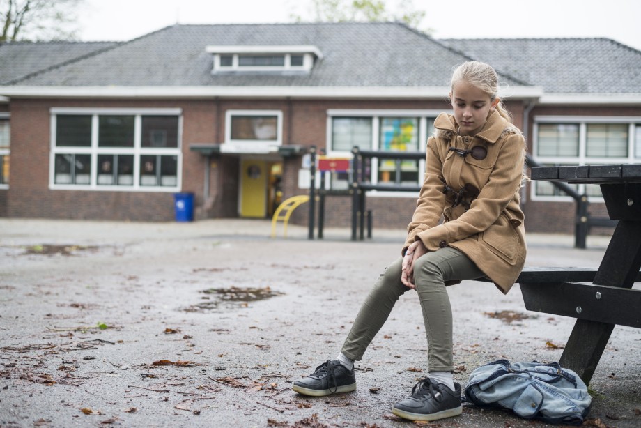 Jente som lener seg inn til bokskap, mens tre jenter står og visker i bakgrunnen. Jente sitter alene i en skolegård.