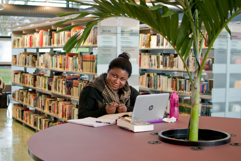 Dame sitter ved rundt bord med palmeplante og studerer på en PC