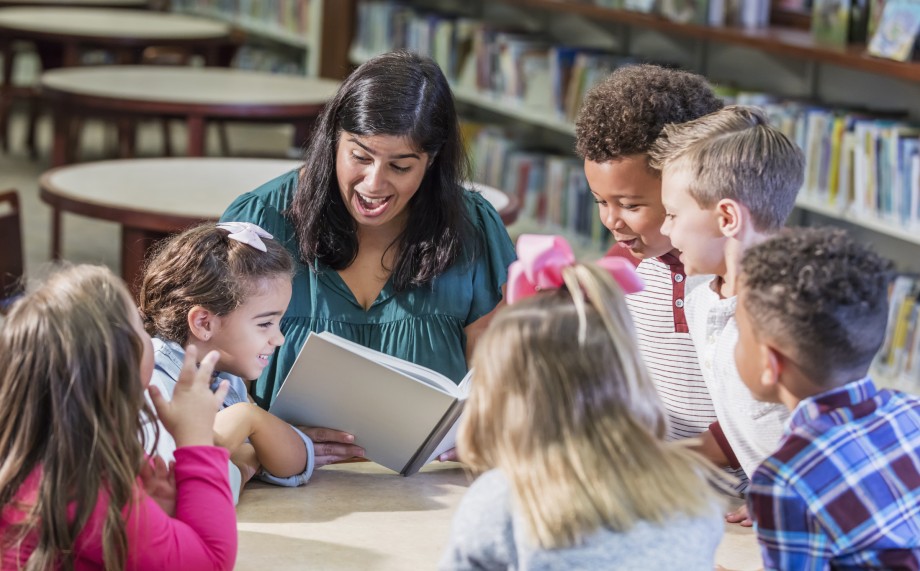 Seks elever i småskolealder og en kvinnelig lærer sitter rundt et bord i et bibliotek. Læreren leser høyt fra en bok og smiler. Elevene lener seg over bordet og er tydelig engasjert i fortellingen.