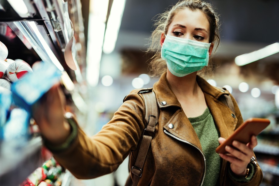 Ung kvinne med ansiktsmaske ved hjelp av mobiltelefon og kjøpe dagligvarer i supermarkedet under viruspandemi. 