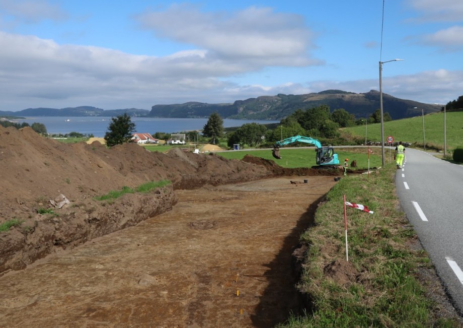 Gule pinner i nedre billedkant markere noen av stolpehullene som kan tilhøre et langhus. Rennesøy ses i bakgrunnen.