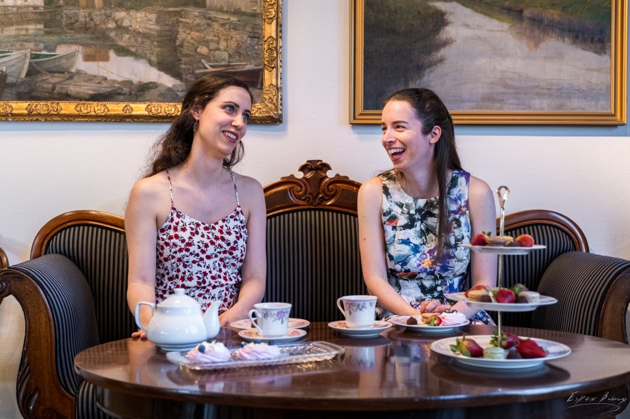 To smilende damer som sitter i en sofa, med et bord foran seg med te og kaker